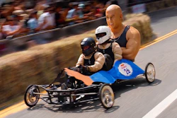 Gravity-powered soapbox cart racing downhill with three drivers in helmets on a street lined with spectators.