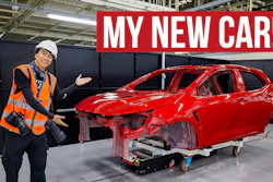Factory worker in safety gear posing next to red car chassis in manufacturing facility