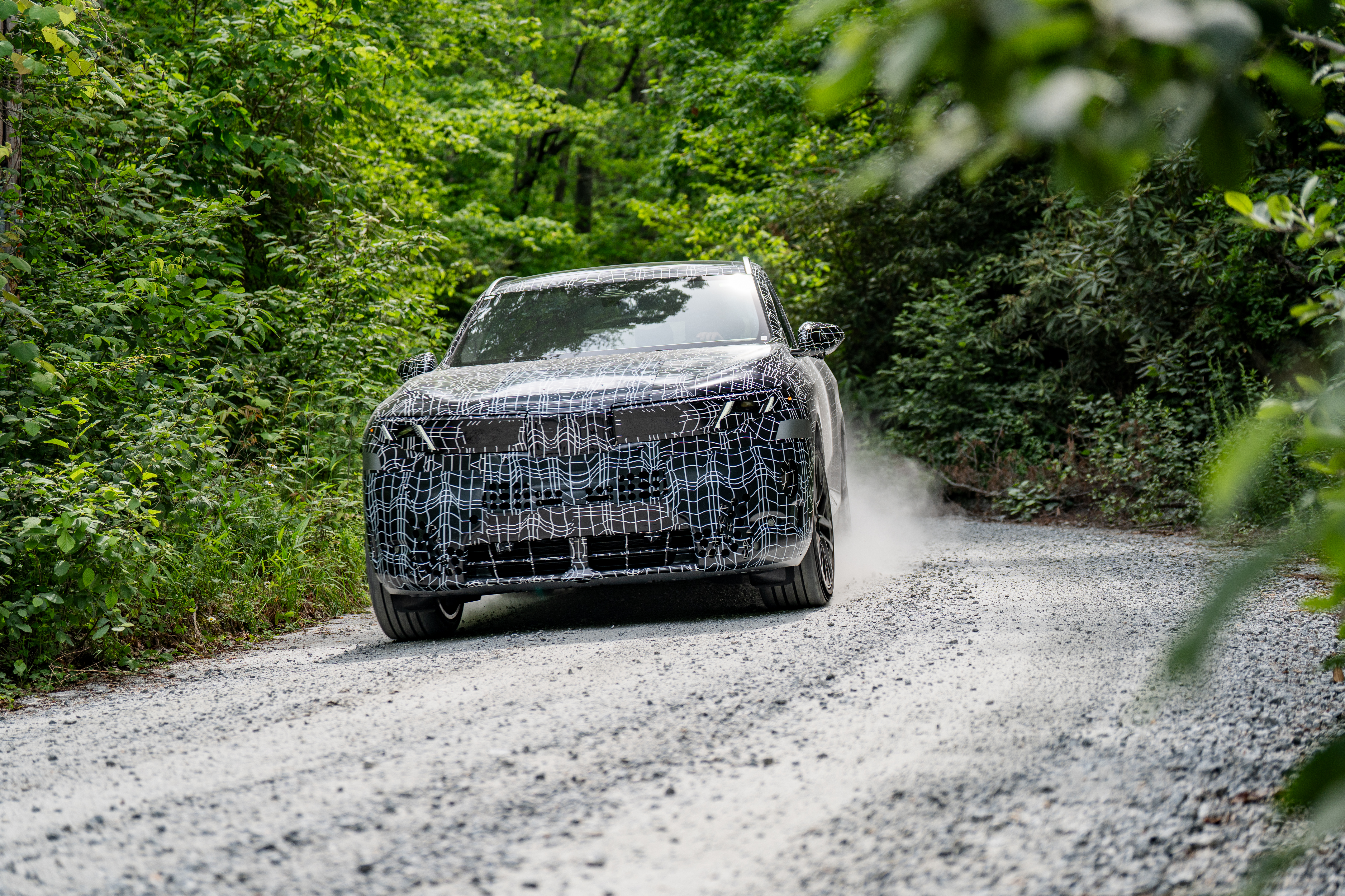 Camouflaged prototype SUV on gravel road surrounded by green forest vegetation during vehicle testing
