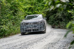 Camouflaged prototype SUV on gravel road surrounded by green forest vegetation during vehicle testing