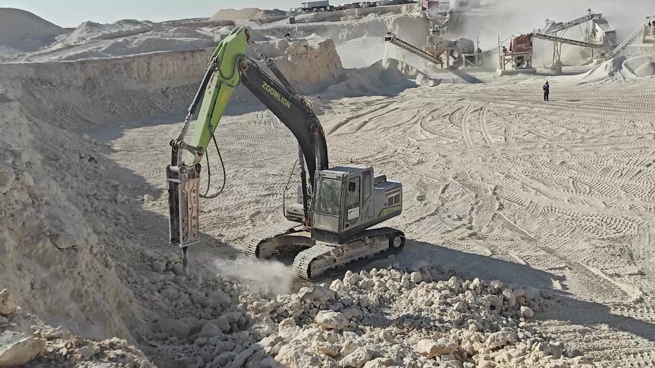 Hydraulic excavator with breaker attachment extracting stone at active quarry site with processing equipment