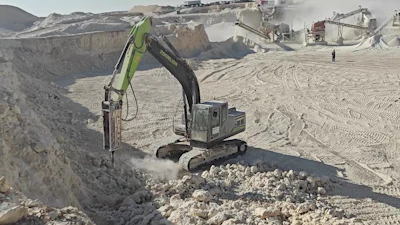 Hydraulic excavator with breaker attachment extracting stone at active quarry site with processing equipment