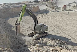 Hydraulic excavator with breaker attachment extracting stone at active quarry site with processing equipment
