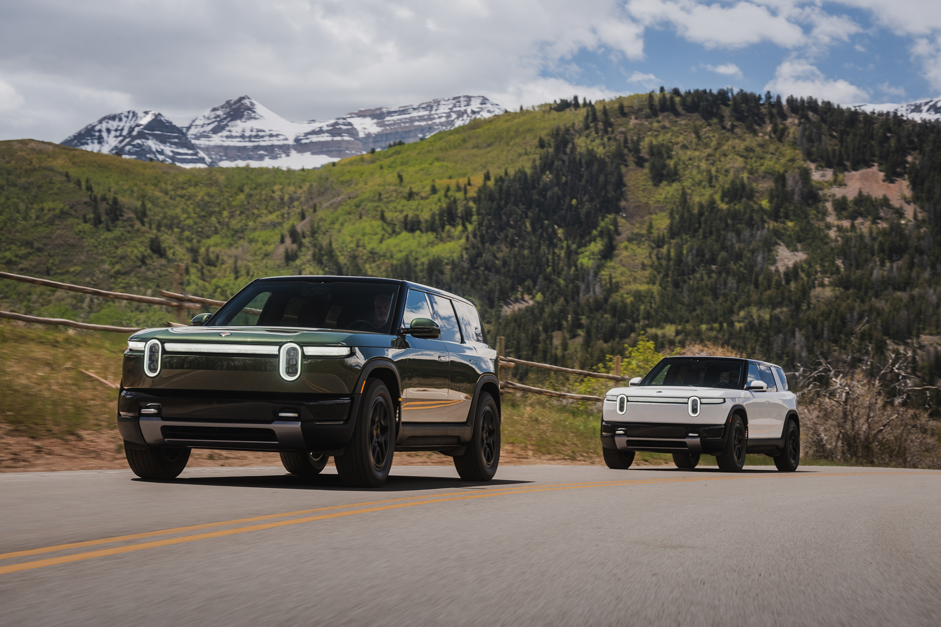 Two Rivian electric vehicles on a mountain road with snow-capped peaks and forested landscape in background