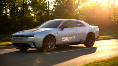 White Dodge Challenger muscle car with Solid State Battery branding driving on a tree-lined road at sunset