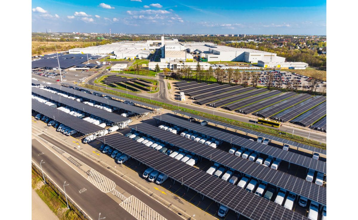 Aerial view of industrial facility with extensive solar panel arrays, white warehouse buildings, and parking areas surrounded by suburban landscape.