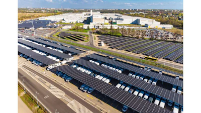 Aerial view of industrial facility with extensive solar panel arrays, white warehouse buildings, and parking areas surrounded by suburban landscape.