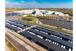 Aerial view of industrial facility with extensive solar panel arrays, white warehouse buildings, and parking areas surrounded by suburban landscape.