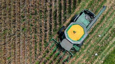 Aerial view of green tractor with yellow grain cart harvesting rows of crops in agricultural field