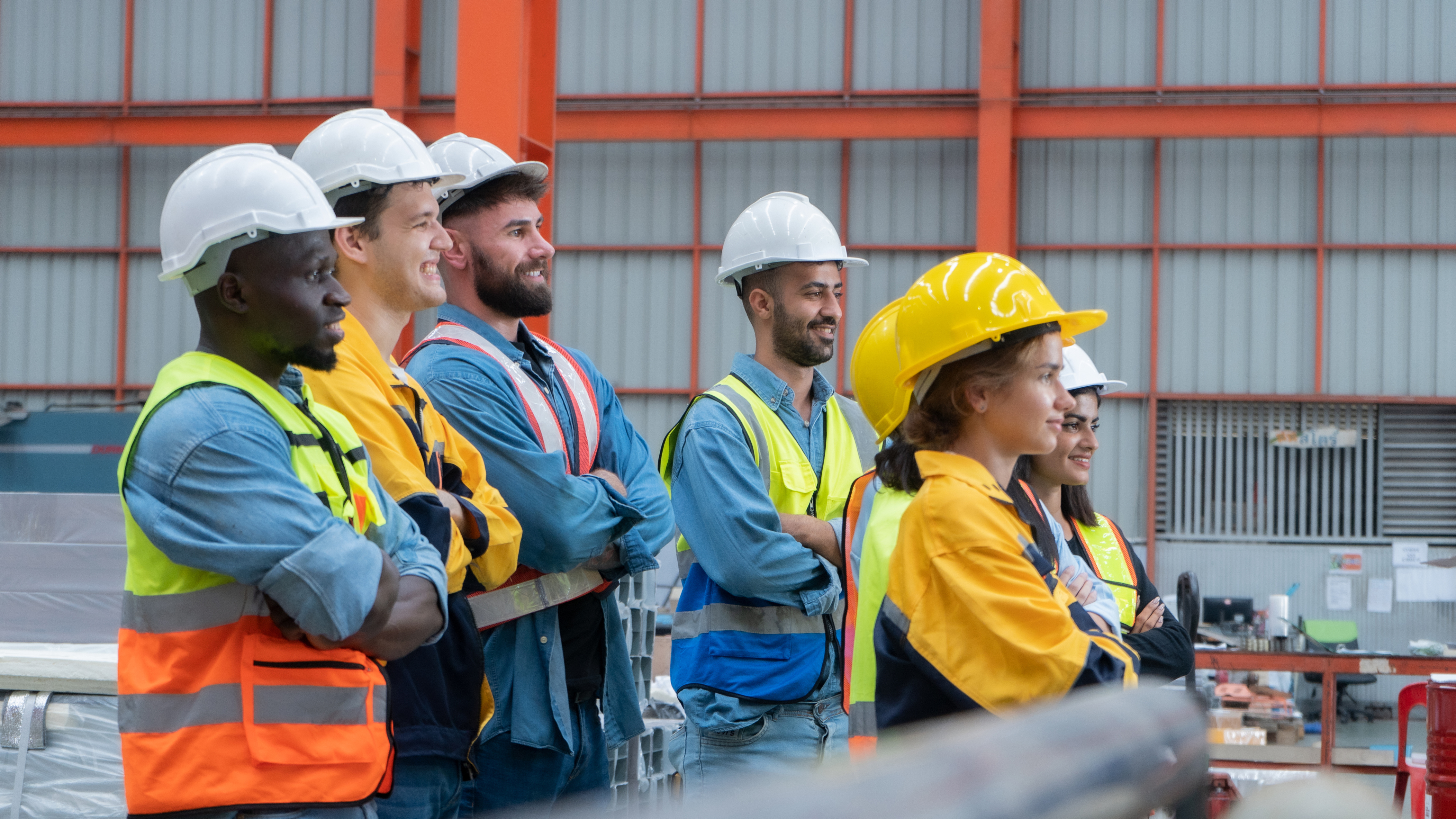 Construction team wearing safety gear standing together at industrial job site with metal building backdrop