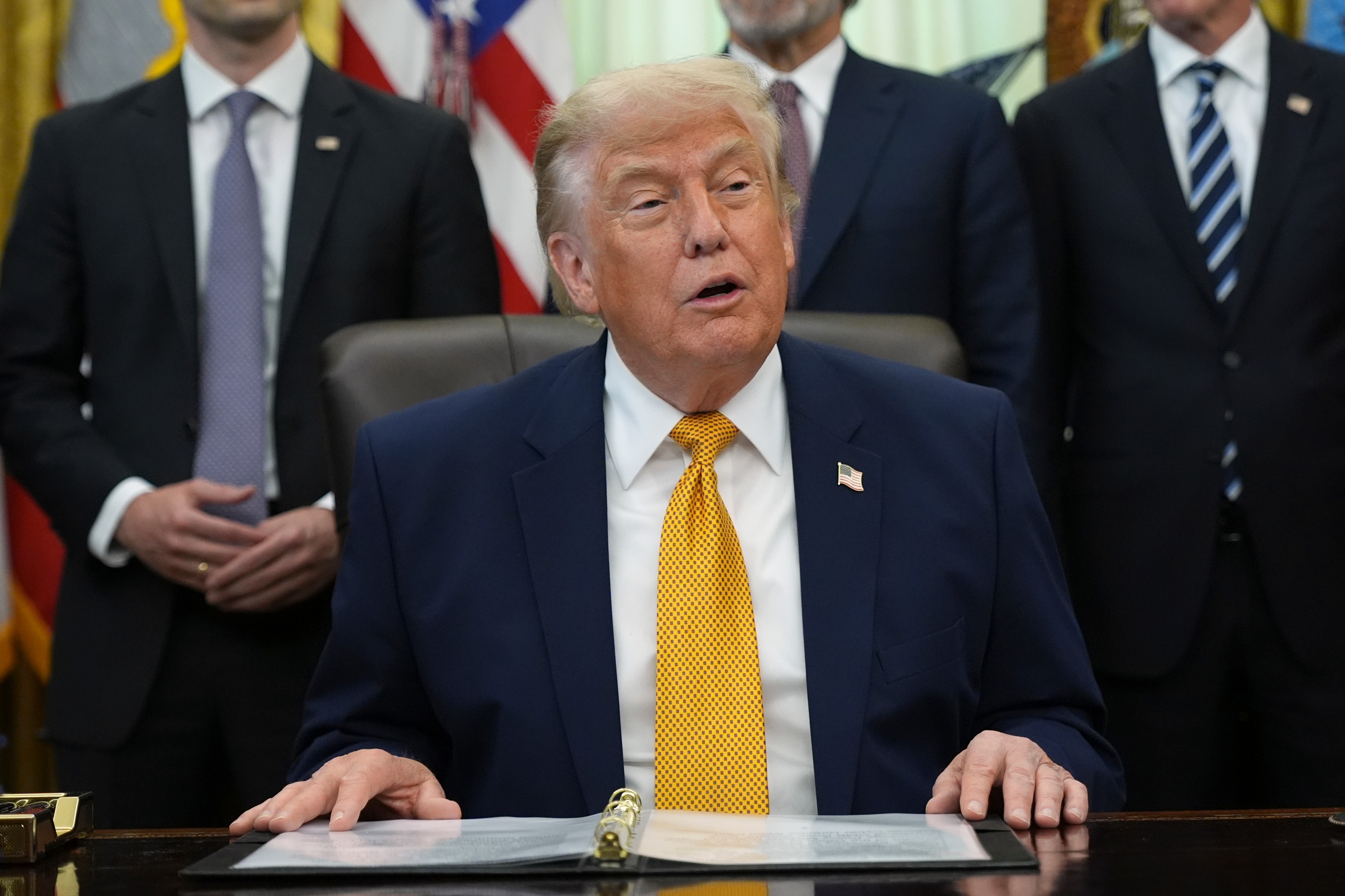 Official seated at Oval Office desk with American flags and staff members in background