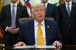 Official seated at Oval Office desk with American flags and staff members in background