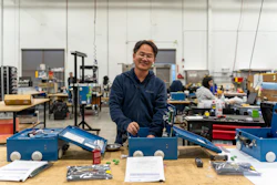 Man in navy jacket stands at workbench with blue toolboxes in a manufacturing workshop facility