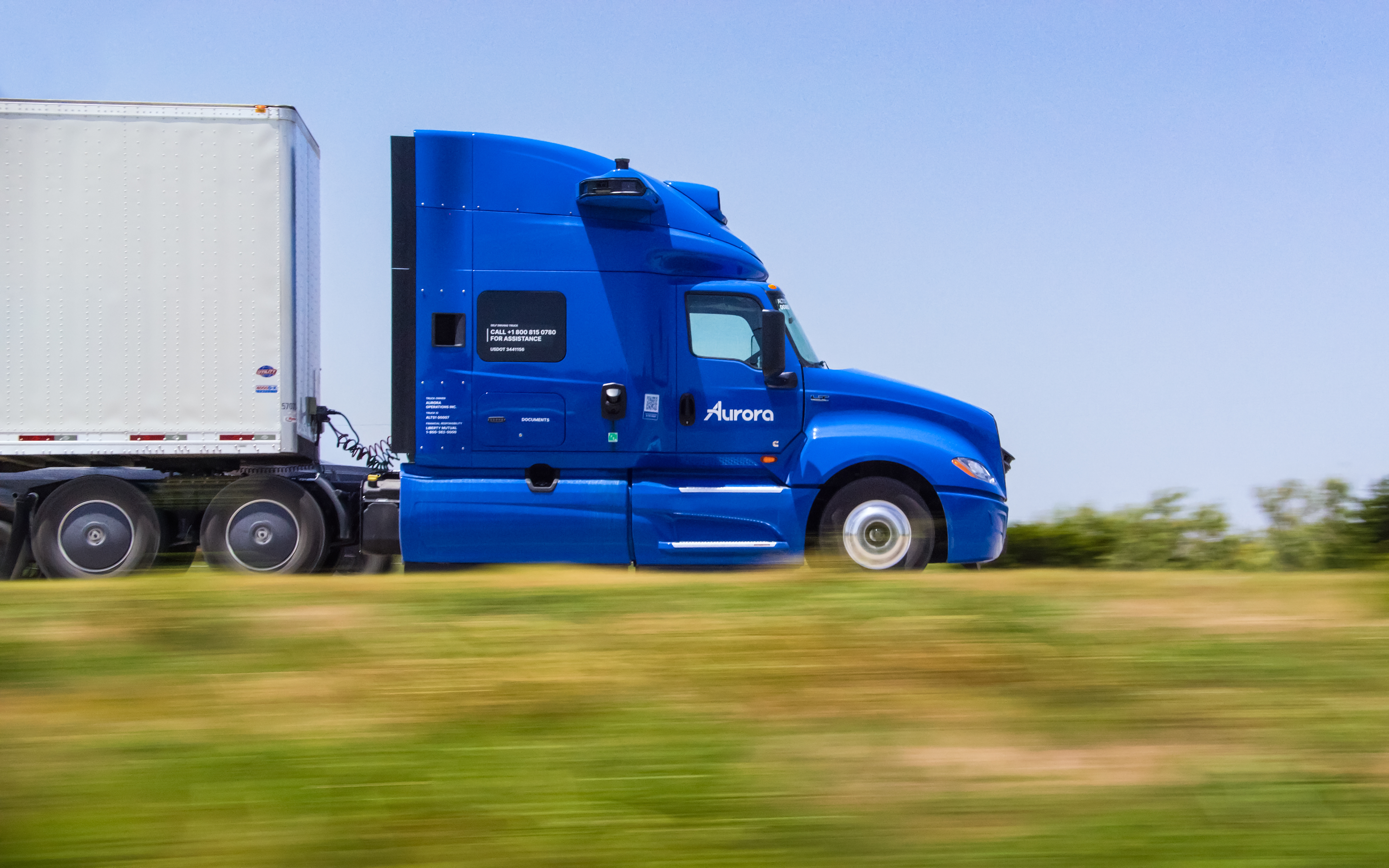 Blue Aurora semi-truck with white refrigerated trailer driving on highway with green fields