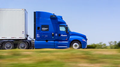 Blue Aurora semi-truck with white refrigerated trailer driving on highway with green fields