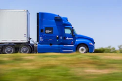 Blue Aurora semi-truck with white refrigerated trailer driving on highway with green fields
