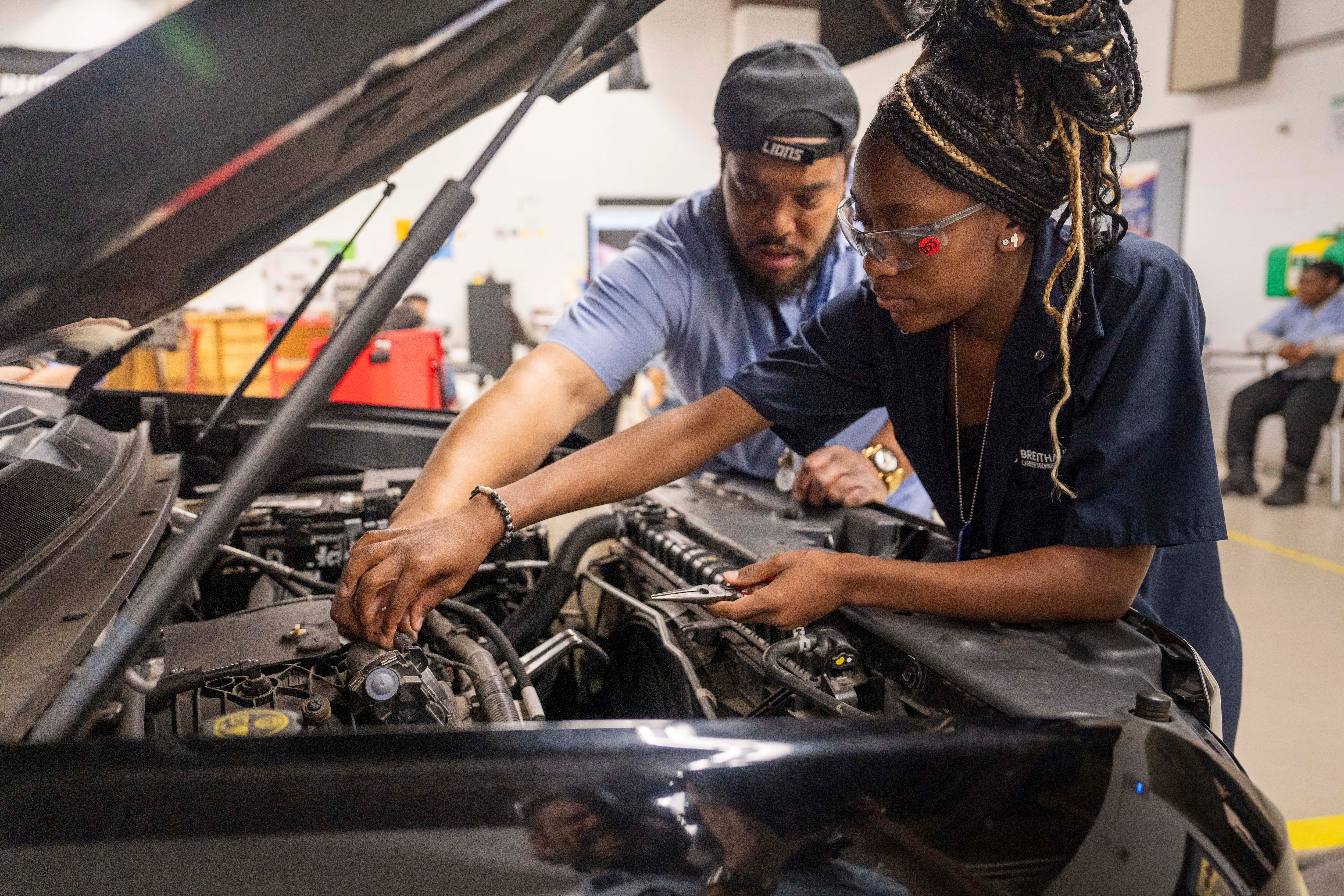 Two automotive technicians inspecting and working on a car engine in a professional workshop facility.