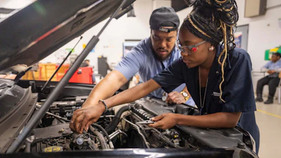 Two automotive technicians inspecting and working on a car engine in a professional workshop facility.