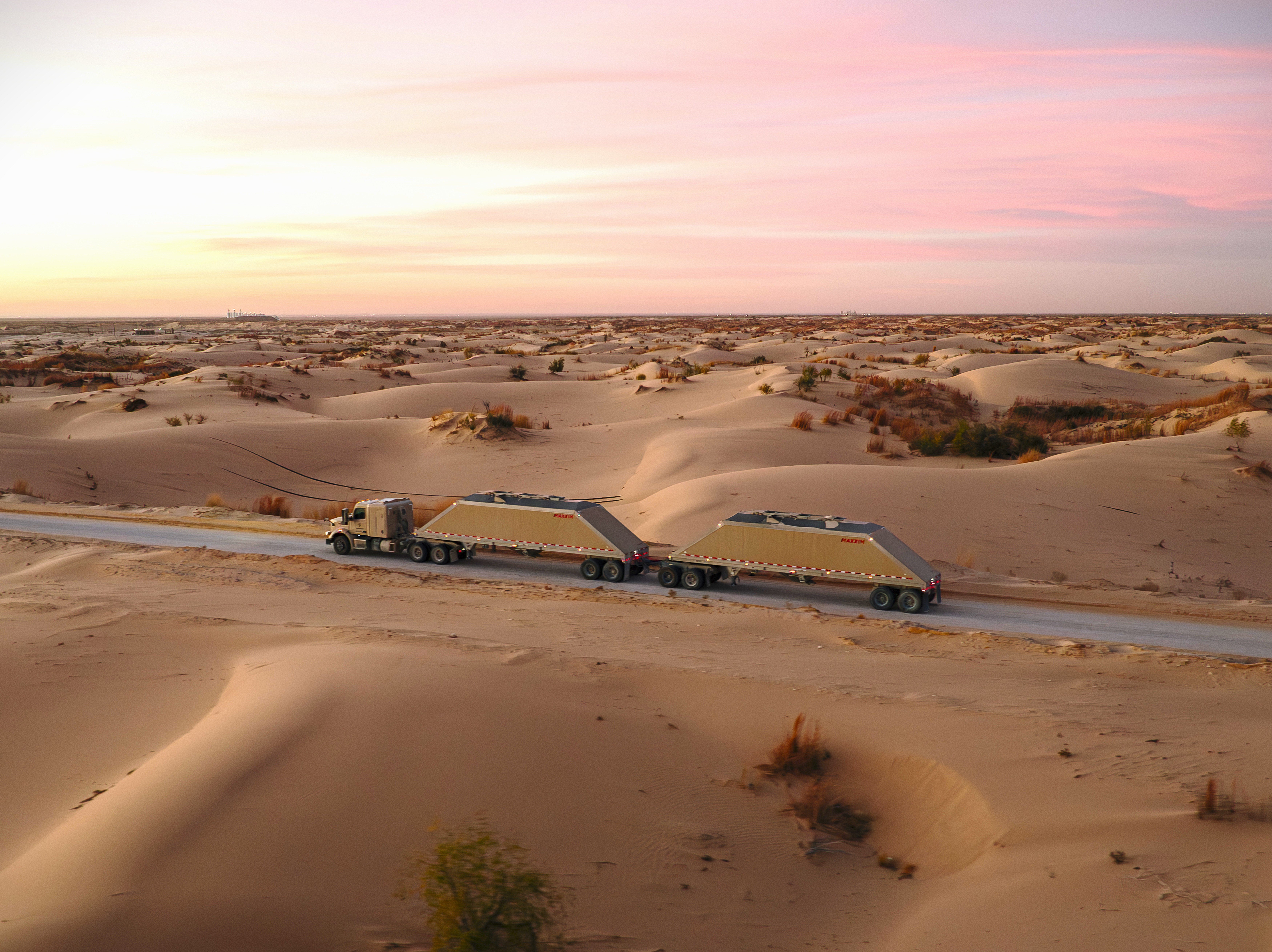 Large cargo truck with trailers traveling through desert landscape at sunset with pink sky and sand dunes