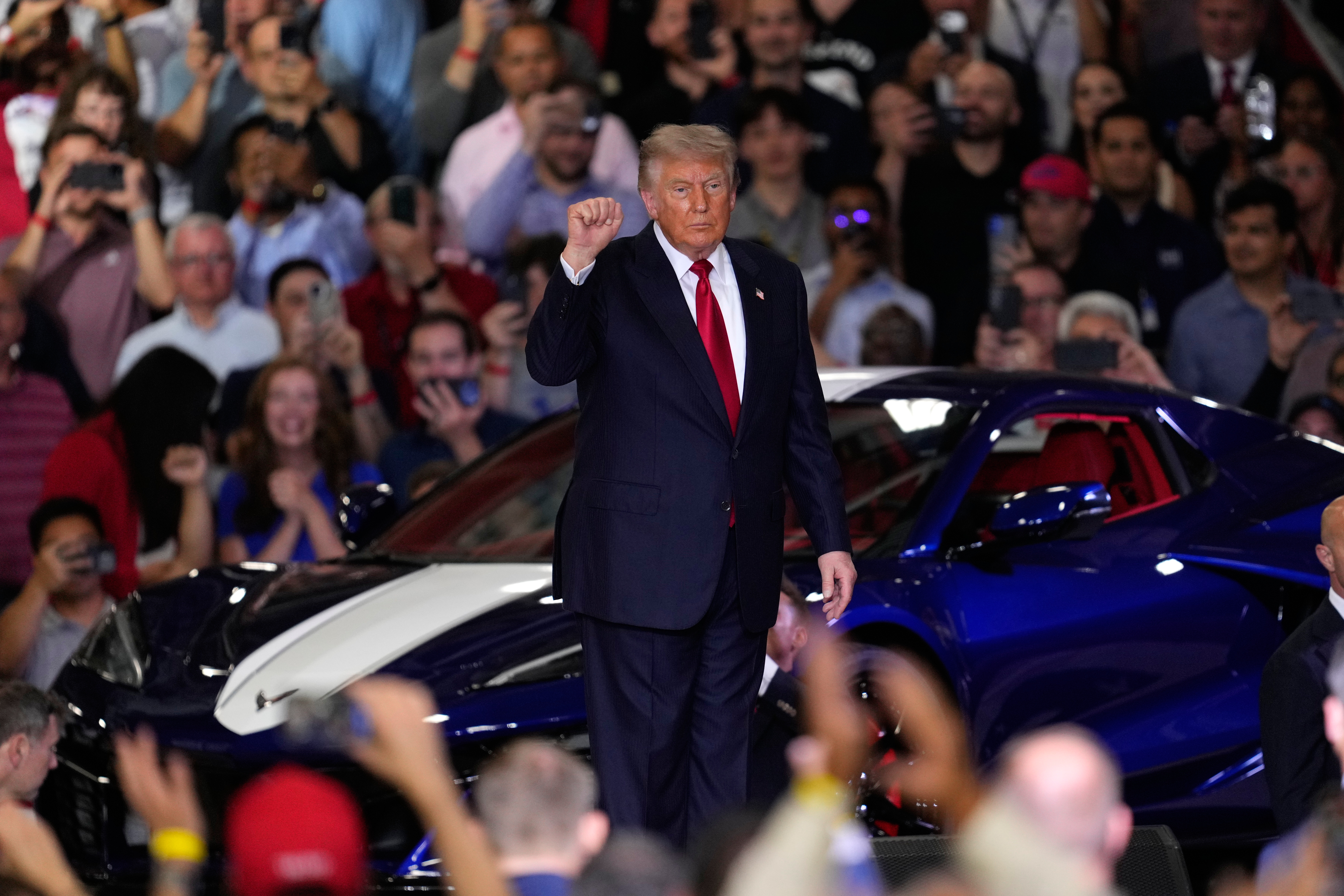 Political rally speaker in dark suit addressing large crowd with blue vehicle visible in background