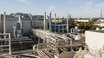 Industrial chemical processing facility with large storage tanks, interconnected piping, and manufacturing equipment under clear sky