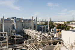 Industrial chemical processing facility with large storage tanks, interconnected piping, and manufacturing equipment under clear sky