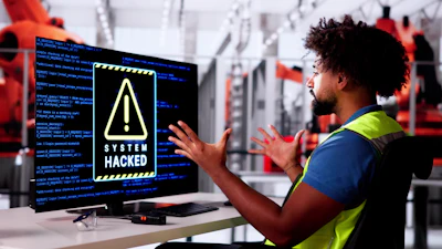 Industrial worker in safety vest gestures at computer monitor displaying 'System Hacked' warning with code in background factory setting