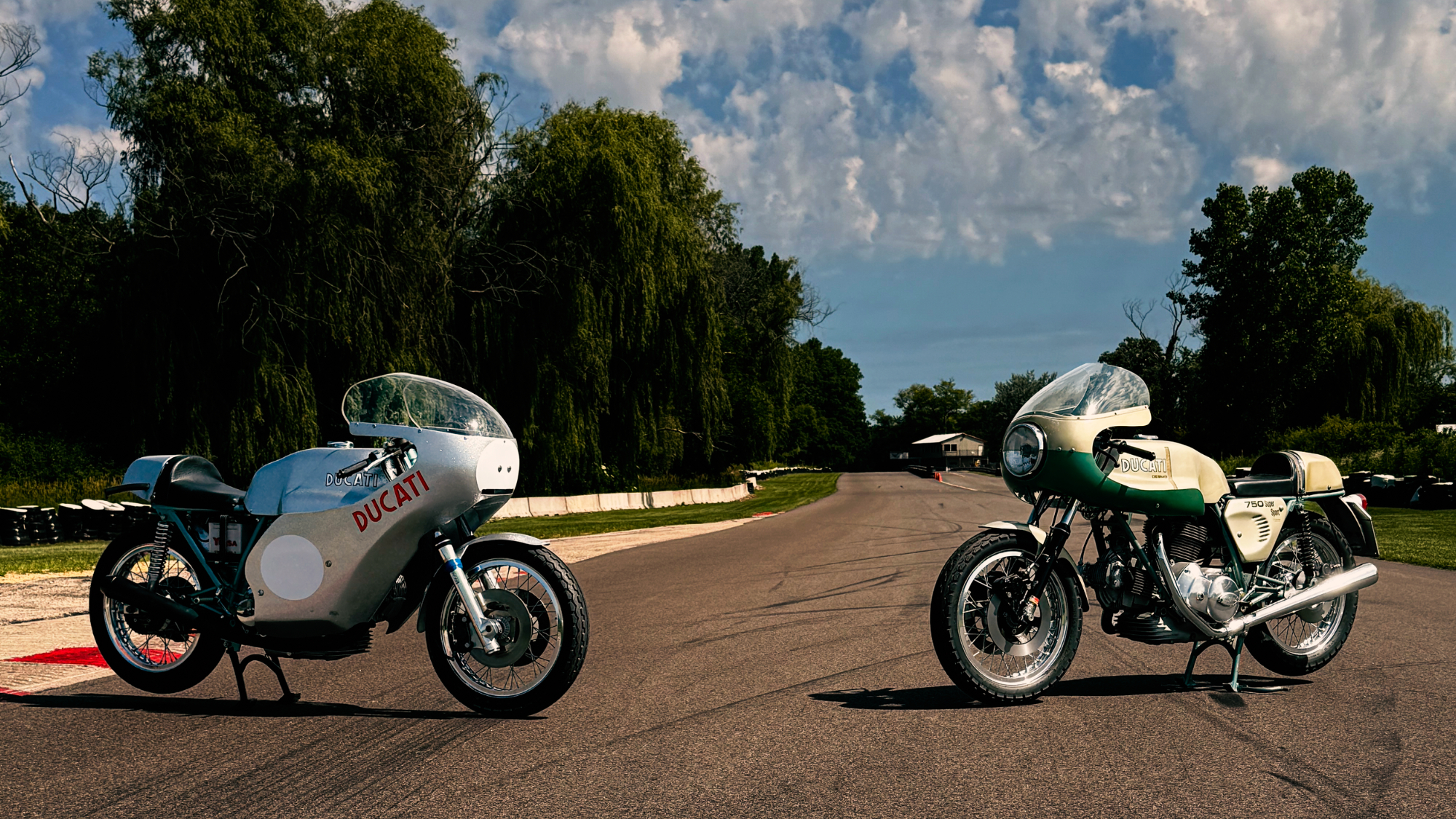 Vintage Ducati and Benelli racing motorcycles parked on a racetrack with trees and sky in background