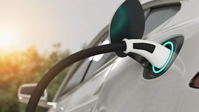 White electric vehicle charging cable connected to car with glowing cyan charging port at sunset