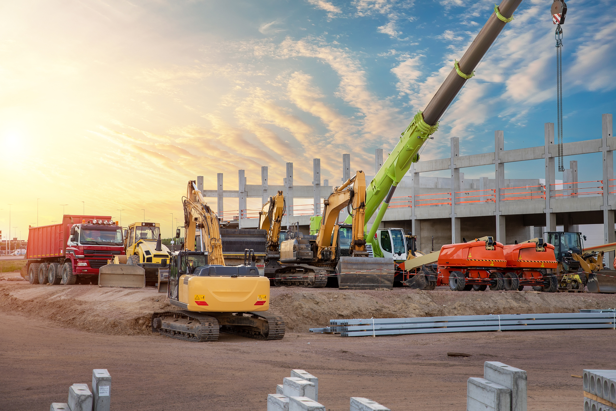 Construction site with excavators, cranes, and heavy machinery at sunset with partially built steel structure.