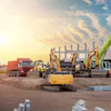 Construction site with excavators, cranes, and heavy machinery at sunset with partially built steel structure.
