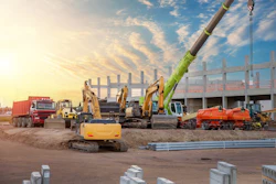 Construction site with excavators, cranes, and heavy machinery at sunset with partially built steel structure.