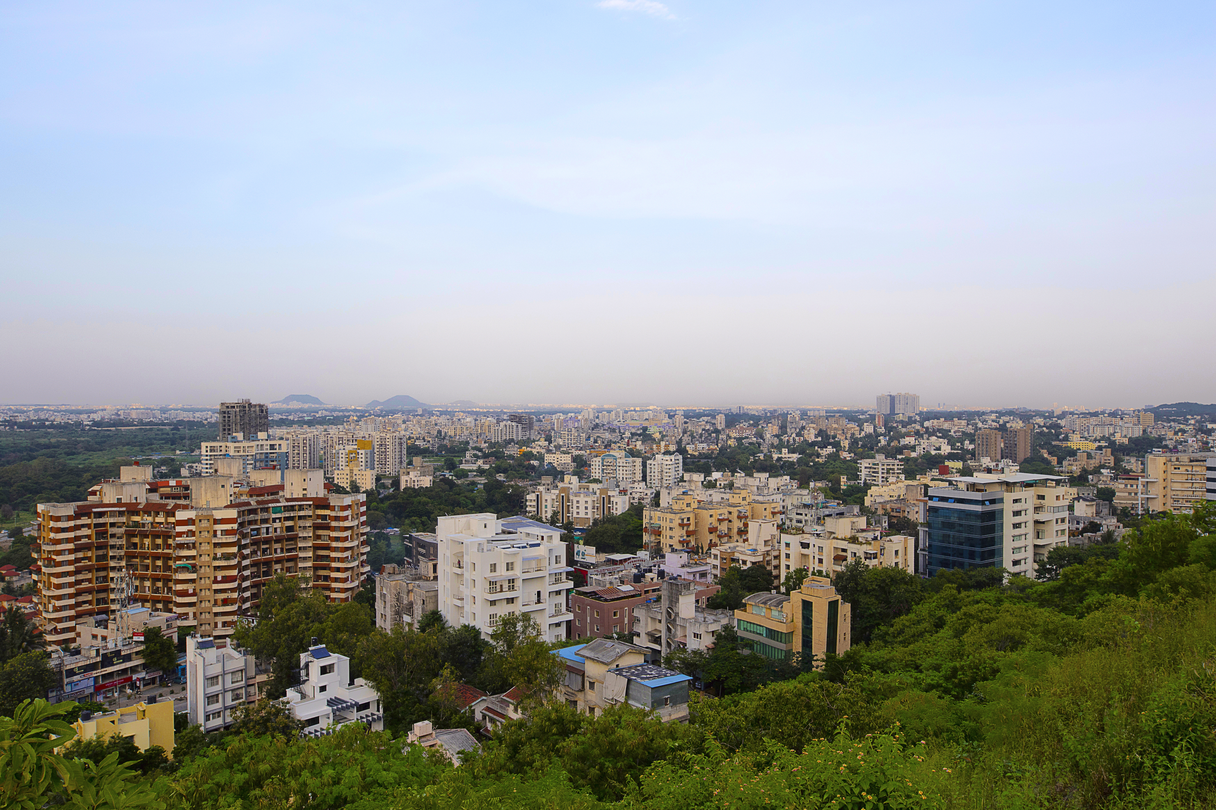 Aerial cityscape with residential and commercial buildings, green vegetation, and distant mountains under hazy sky