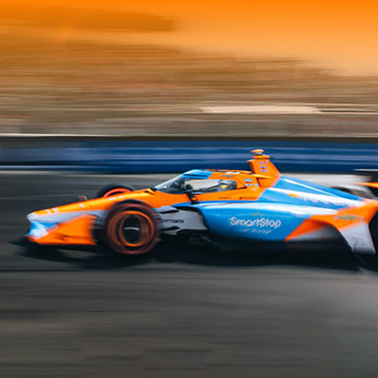 Orange and blue IndyCar racing vehicle in motion on track at sunset