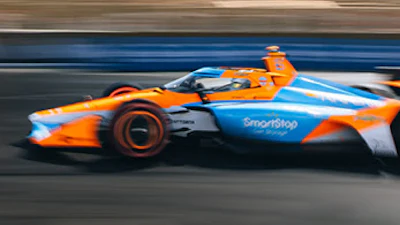 Orange and blue IndyCar racing vehicle in motion on track at sunset