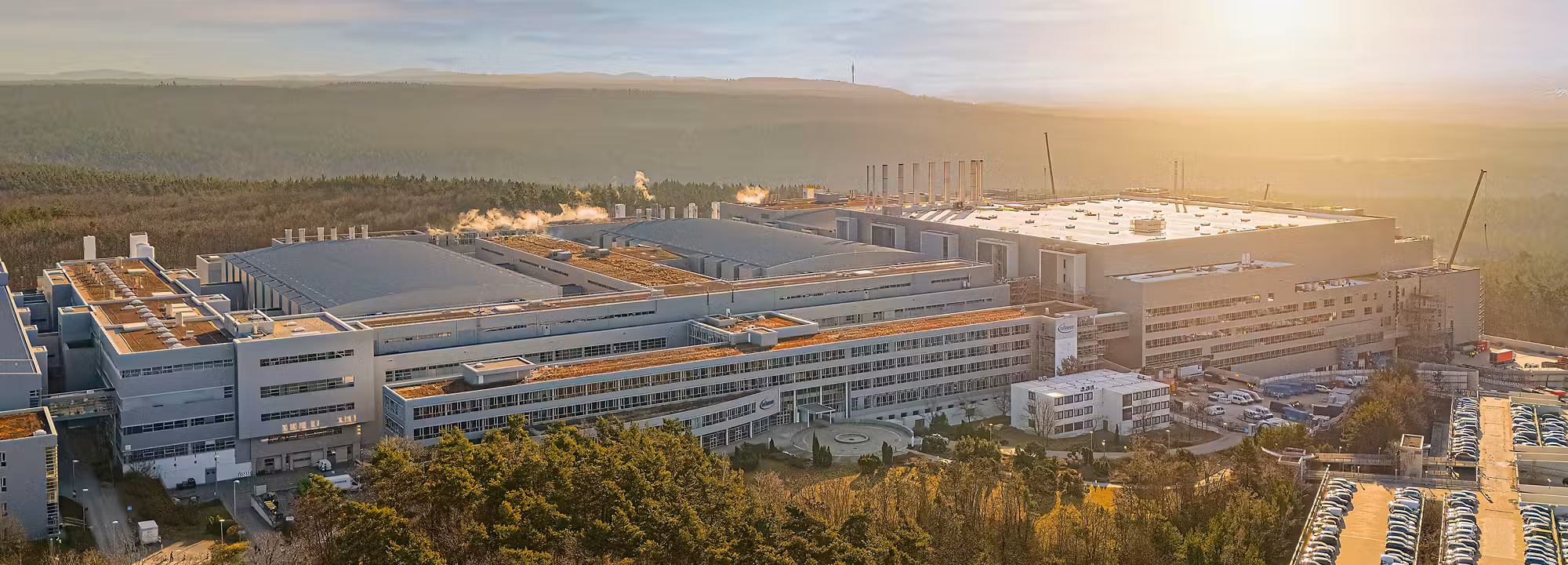 Aerial view of large industrial manufacturing complex with multi-story buildings, solar panels, and parking areas surrounded by forest at sunset.