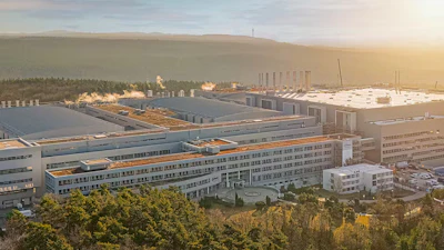 Aerial view of large industrial manufacturing complex with multi-story buildings, solar panels, and parking areas surrounded by forest at sunset.