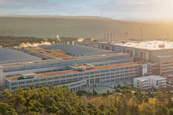 Aerial view of large industrial manufacturing complex with multi-story buildings, solar panels, and parking areas surrounded by forest at sunset.