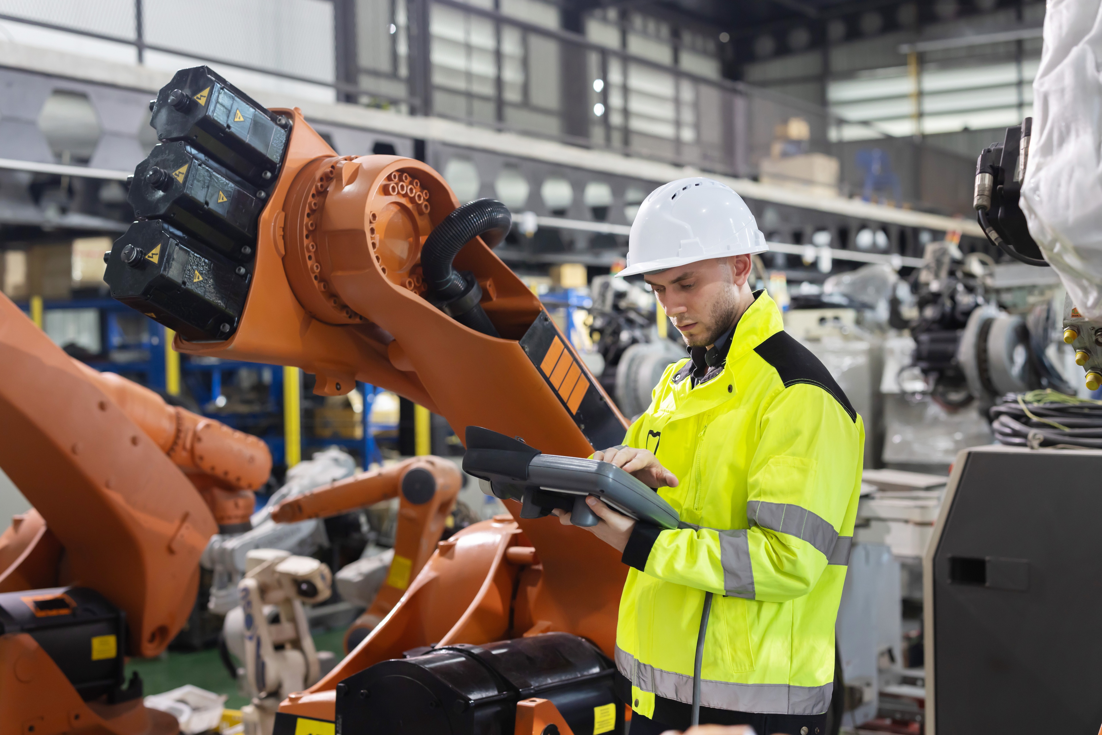 Factory worker in safety gear operating an orange industrial robotic arm in a manufacturing facility