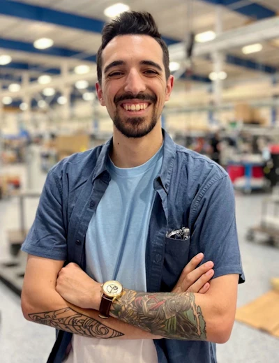 Smiling man with dark hair and beard wearing blue shirt and watch, standing in industrial warehouse setting