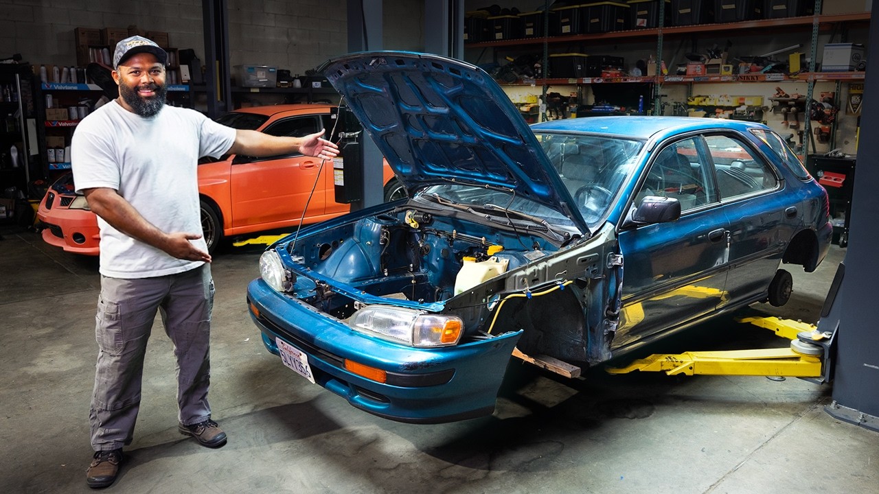 Mechanic in workshop posing with blue car engine and orange vehicle in background