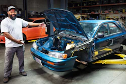 Mechanic in workshop posing with blue car engine and orange vehicle in background