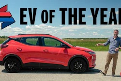Red electric SUV with 'EV of the Year' text and man presenting vehicle in outdoor field setting