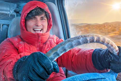 Young person wearing bright red winter jacket and hood smiling while holding steering wheel in snowy landscape