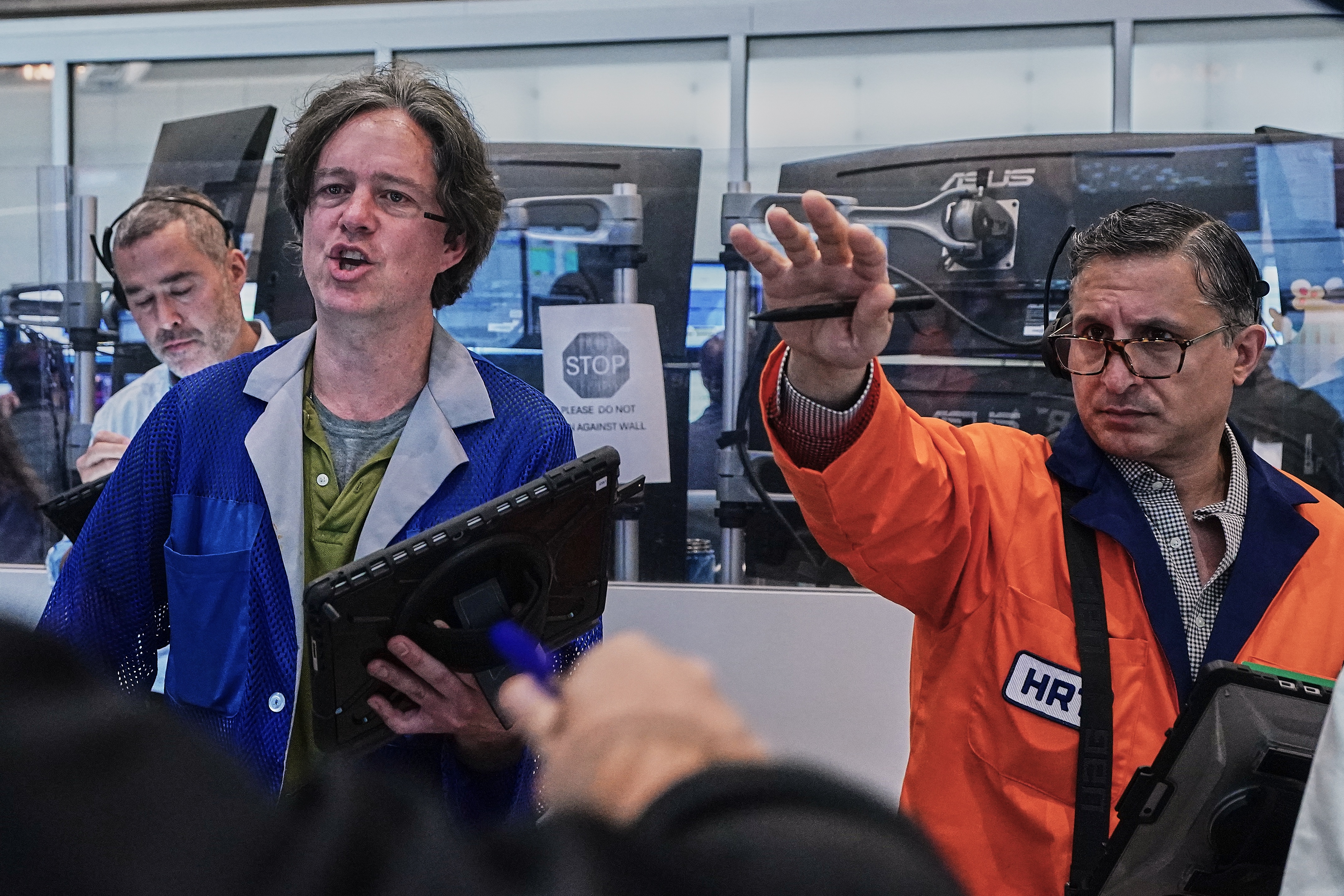 Three men demonstrate technology at an industrial event, with one wearing an HR badge and orange safety vest gesturing toward computer equipment.