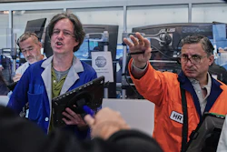 Three men demonstrate technology at an industrial event, with one wearing an HR badge and orange safety vest gesturing toward computer equipment.