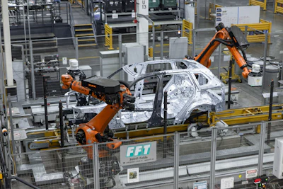 Robotic arms assembling white car body in automotive manufacturing facility with yellow safety barriers