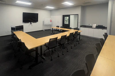 Modern conference room with wooden tables, black chairs, wall-mounted TV, and whiteboard in professional office setting