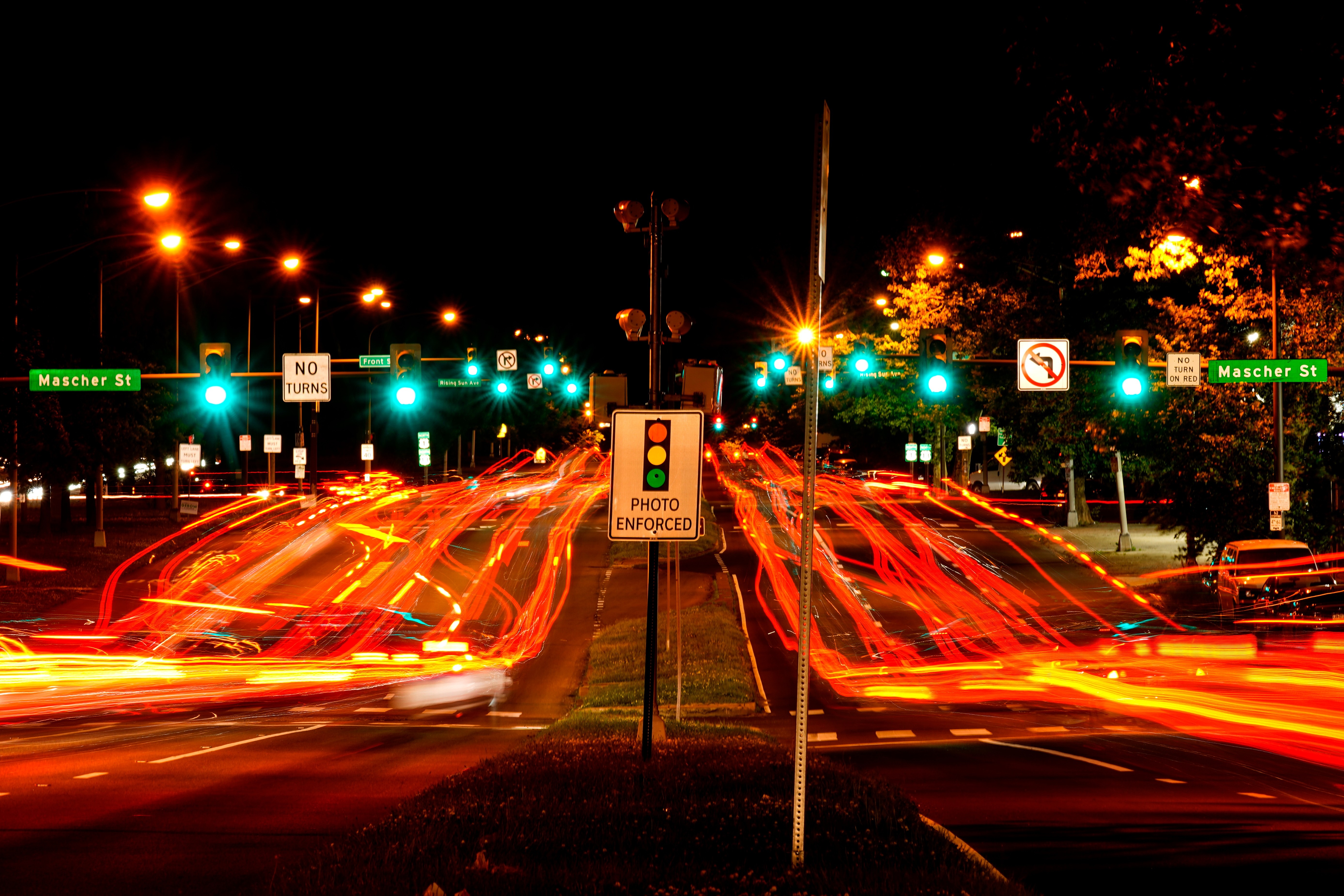 Night traffic intersection with red vehicle light trails, illuminated street signs, and photo enforcement sign at urban crossroads