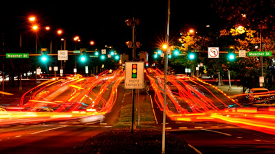 Night traffic intersection with red vehicle light trails, illuminated street signs, and photo enforcement sign at urban crossroads